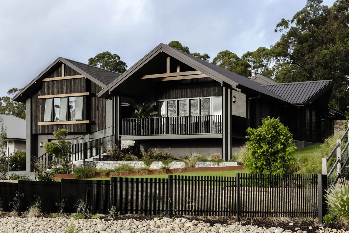 Modern dark timber-clad house with gabled roof, elevated deck, black railing, and landscaped front yard with stone edging and native plants.